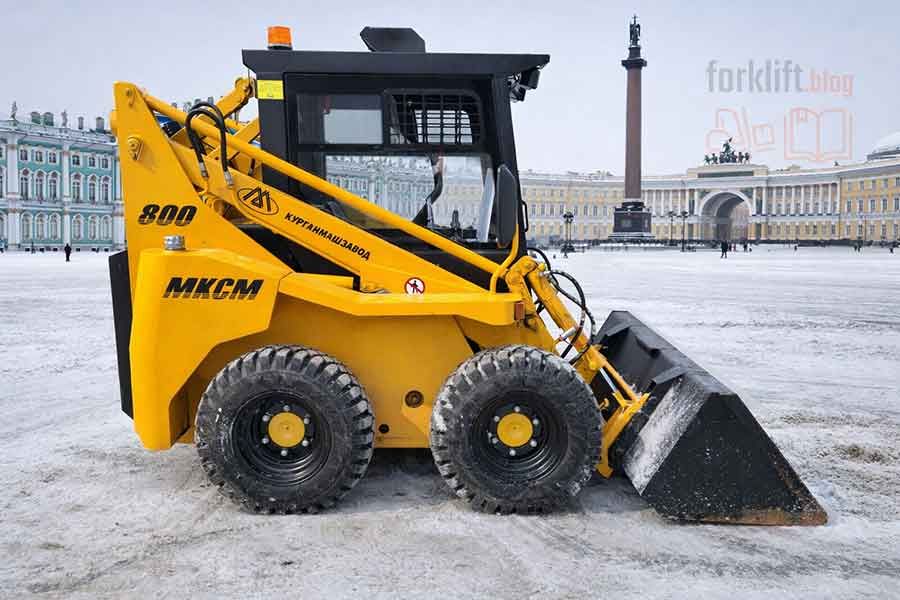Жёлтый мини-погрузчик МКСМ с ковшом очищает заснеженную городскую территорию, надёжное решение для эксплуатации в условиях сильных морозов и интенсивных осадков.